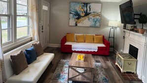 A living room with a red sofa, white sofa, and a coffee table with a fruit basket. There is a painting on the wall, a television, and a fireplace. - Cozy & Quite 2BR 1Bath Duplex Private Entrance Perfect for work Stays (Valdosta)