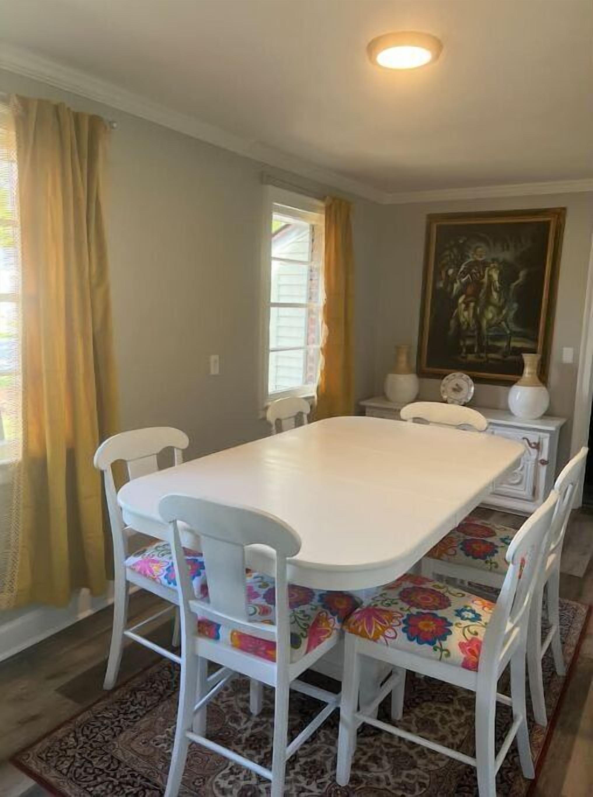 A dining area with a white table and chairs, a patterned rug, a painting on the wall, and a window with curtains.