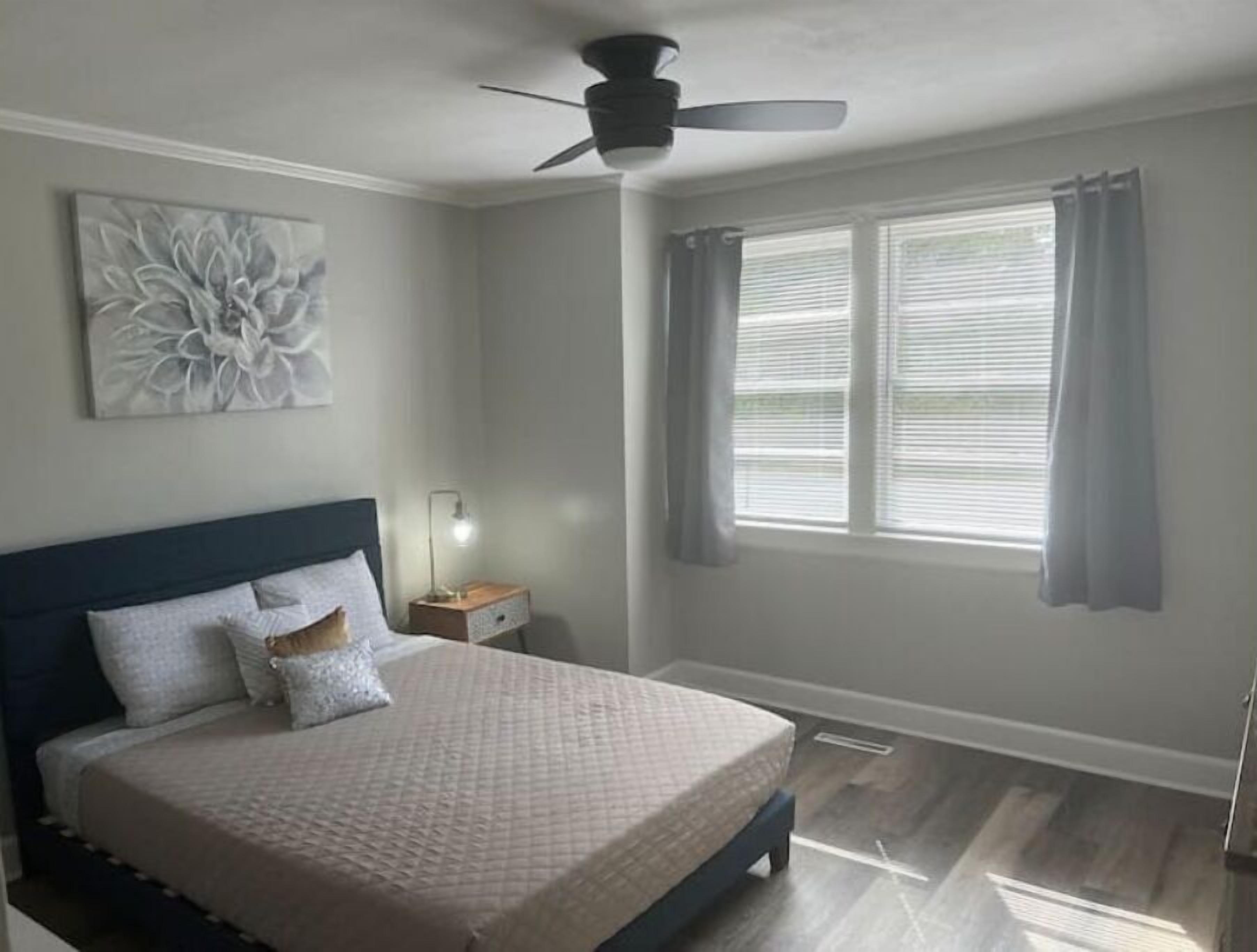 Quarto com cama, ventoinha no teto, mesa de cabeceira com abajur e janela com cortinas.