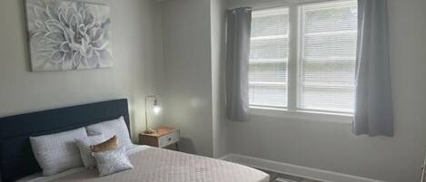 Quarto com cama, ventoinha no teto, mesa de cabeceira com abajur e janela com cortinas.