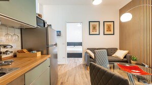 Ett modernt kök med kylskåp, mikrovågsugn och en bänkskiva i trä. Det finns en matplats med ett glasbord och stolar, och ett vardagsrum med en soffa och en TV.