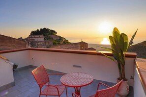 Eine Dachterrasse mit Tisch und Stühlen, einem Blumentopf und Blick auf eine Hügelstadt bei Sonnenuntergang.