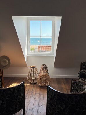 A room with a sloped ceiling, wooden flooring, and a large window overlooking a beach. There are two wicker lamps and a hat on the floor. - Beautiful Seaside Residence - ADELOUANE (Les Sables-d'Olonne)