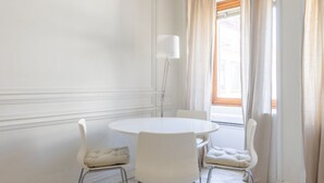 A room with a round white table, four white chairs, a floor lamp, and a window with curtains.