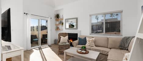 Una sala moderna con sofá, sillón y mesa de centro. Hay una chimenea, un televisor y un ventanal corredera que da a un área exterior.