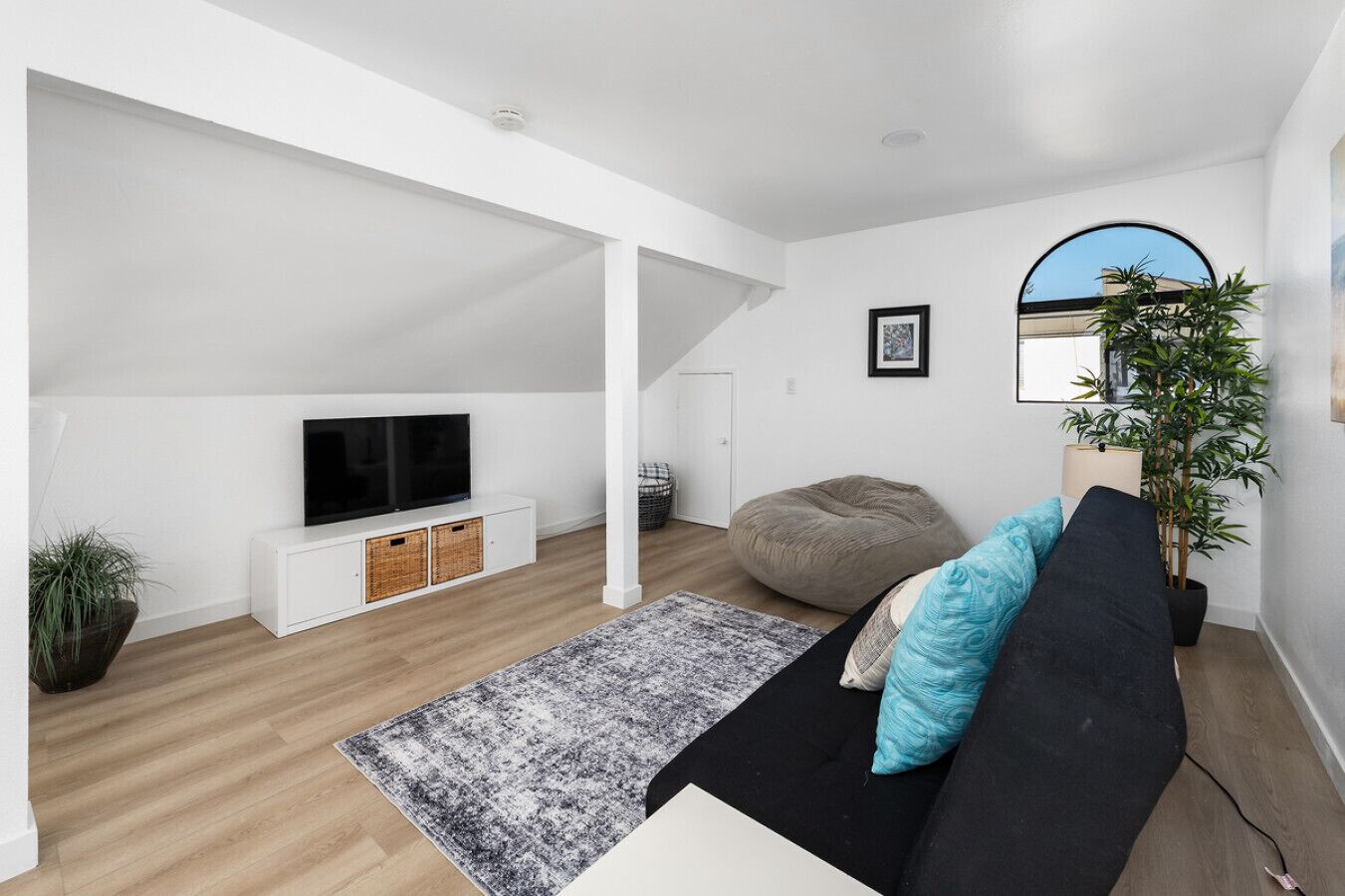 A modern living room with a black sofa, a grey patterned rug, a television on a white cabinet, a large potted plant, and a staircase leading to an upper level.