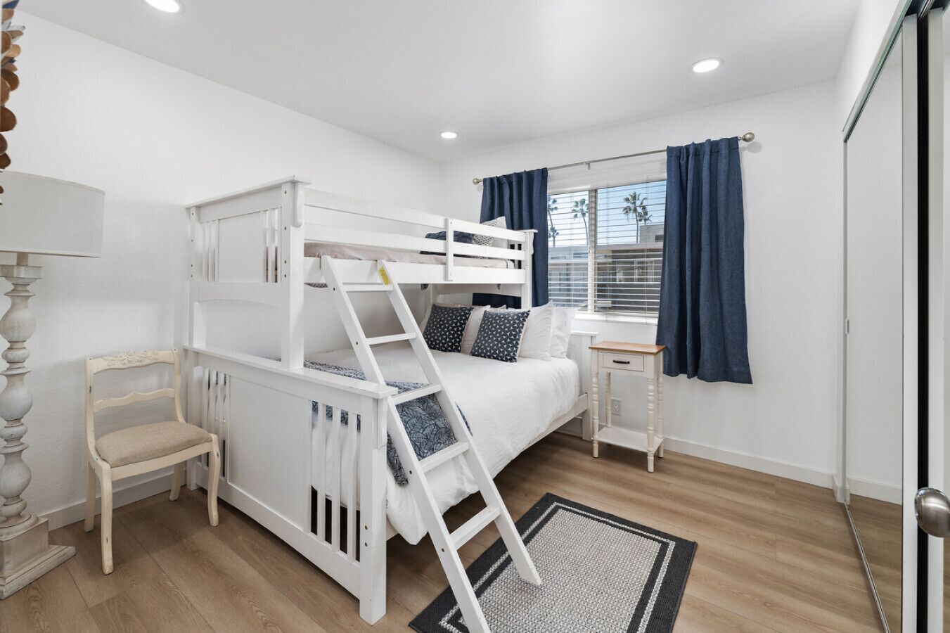 Een slaapkamer met een stapelbed, een klein nachtkastje, een stoel en een raam met gordijnen.
