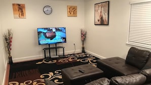 A living room with a leather sofa, a flat-screen TV mounted on the wall, a clock, and two vases with flowers. - Room in House - The Modern Gem Furnished Bedroom (Alexandria)