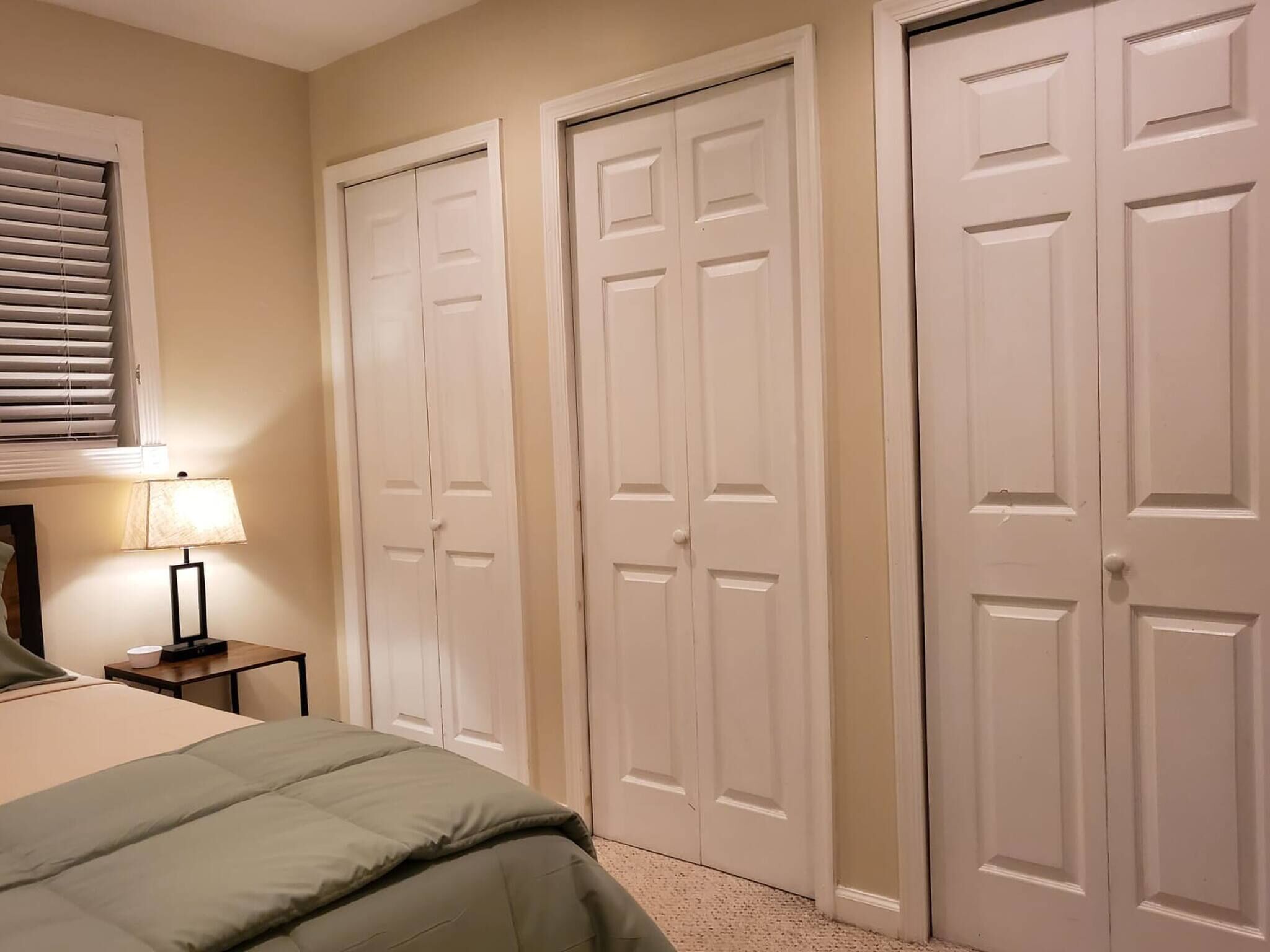 A bedroom with a bed, a nightstand, a lamp, and three white doors.