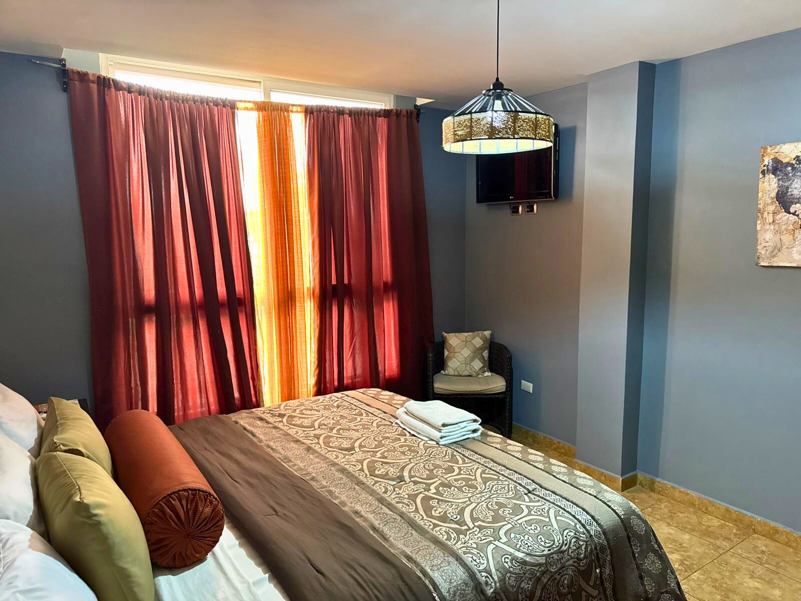 Un dormitorio con cama, cortinas, una silla, una lámpara colgante y una obra de arte en la pared.