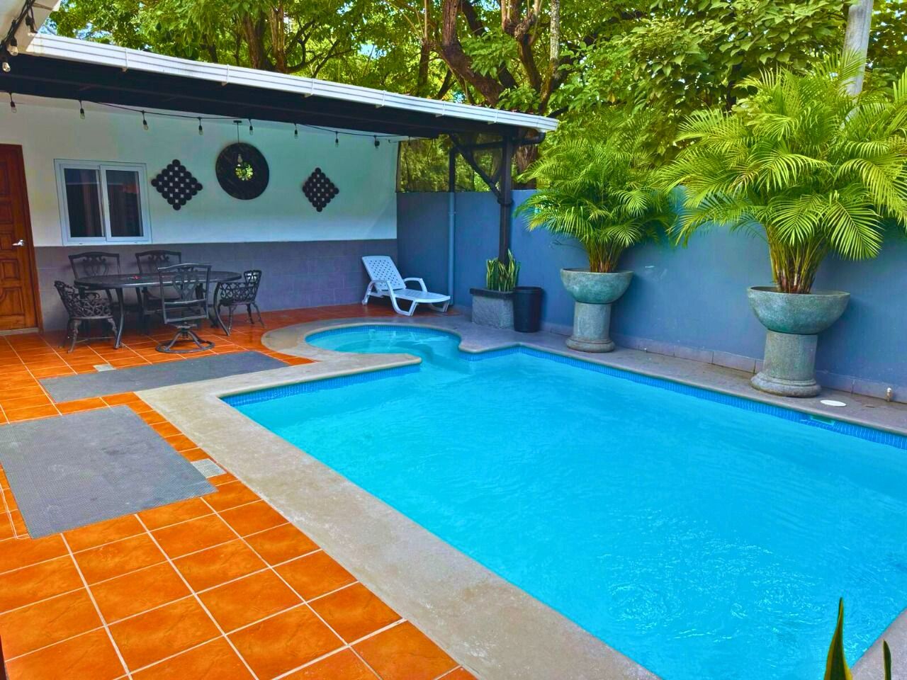 A swimming pool with a tiled deck, a covered seating area, and potted plants.