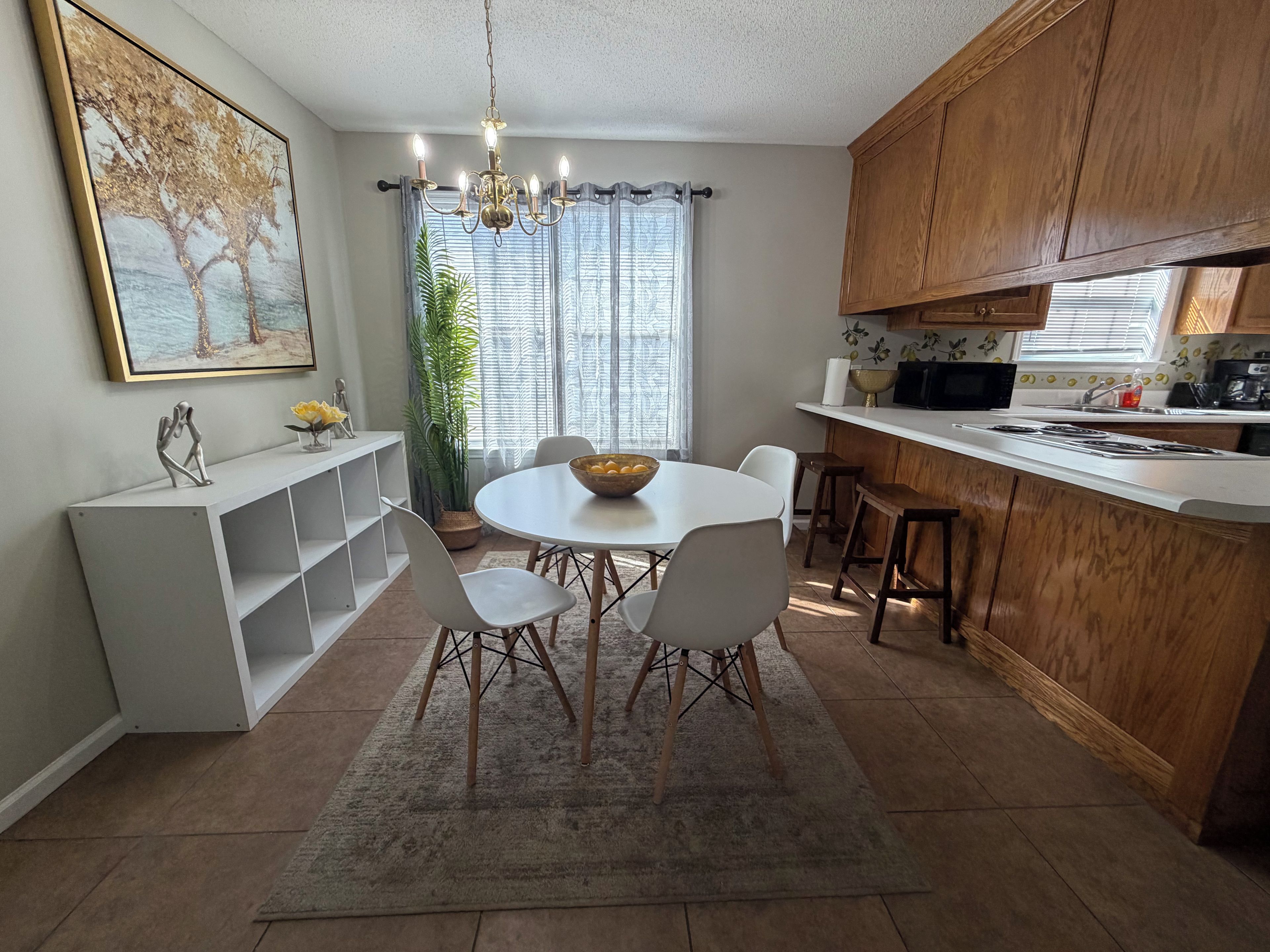 A modern kitchen with a dining area, featuring a chandelier, wooden cabinets, and a large window with curtains.