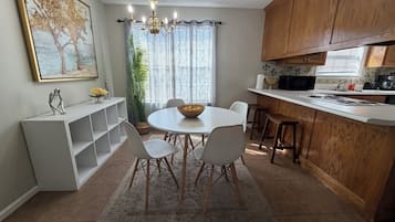 A modern kitchen with a dining area, featuring a chandelier, wooden cabinets, and a large window with curtains.