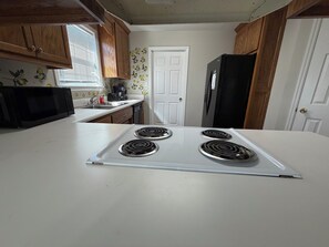 A kitchen with a white countertop, a stove with four burners, wooden cabinets, a black microwave, and a refrigerator. - Peaceful 3BR Home Near Moody AFB + Parking (Valdosta)