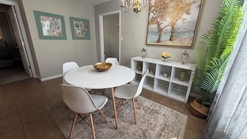 Un coin repas avec une table ronde, des chaises blanches, un lustre, un tableau et une plante en pot.