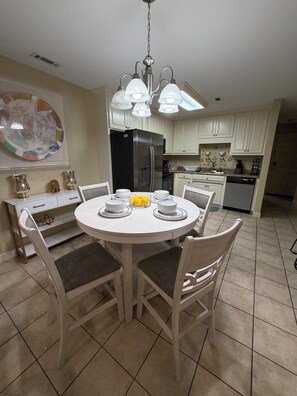 A dining area with a round table, chairs, a chandelier, and a refrigerator in the background.