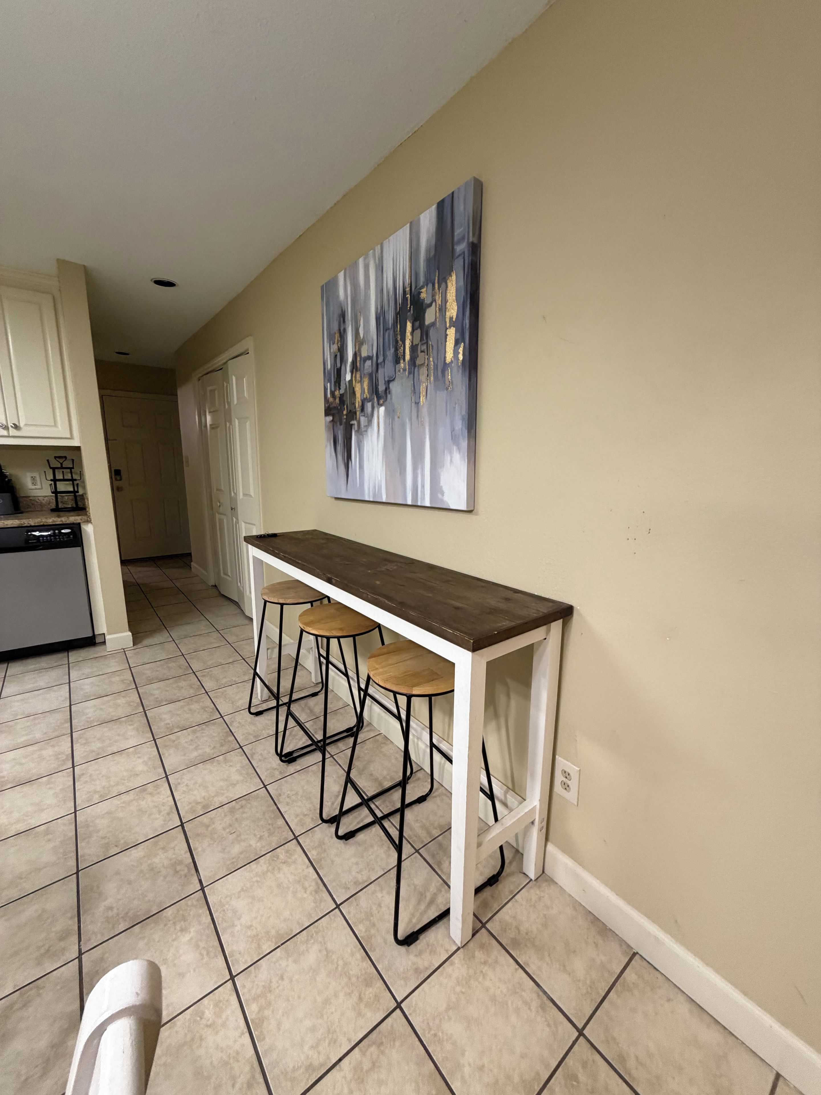 A kitchen with a white countertop, three bar stools, and a painting on the wall.
