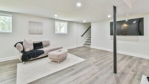 Un salon spacieux avec un canapé de couleur claire, un pouf assorti et un grand téléviseur à écran plat fixé au mur.