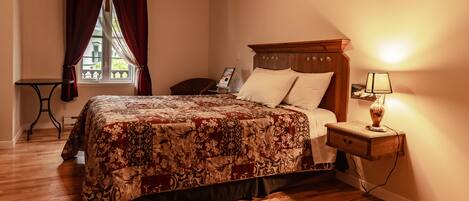 Un dormitorio con una cama de madera, una mesita de noche con lámpara, una ventana con cortinas y una pequeña mesa.