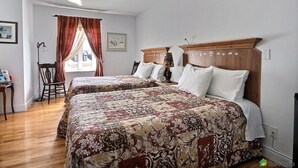 A hotel room with two beds, a ceiling fan, a window with curtains, a chair, and a small table with a lamp. - Auberge Akamaraska (Kamouraska)