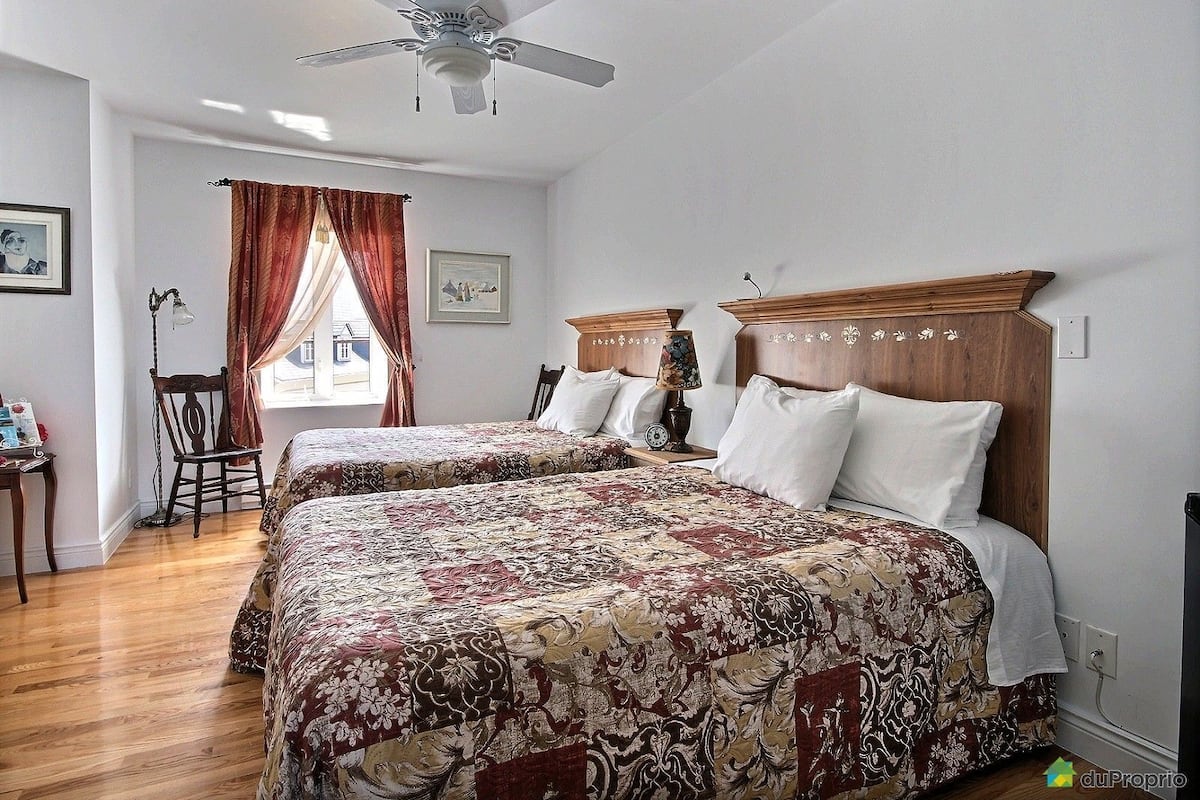 A hotel room with two beds, a ceiling fan, a window with curtains, a chair, and a small table with a lamp.