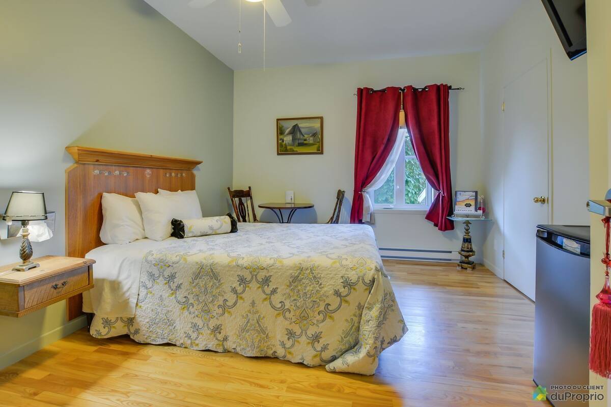 A bedroom with a bed, a nightstand, a desk, a chair, a ceiling fan, a window with red curtains, and a painting on the wall.