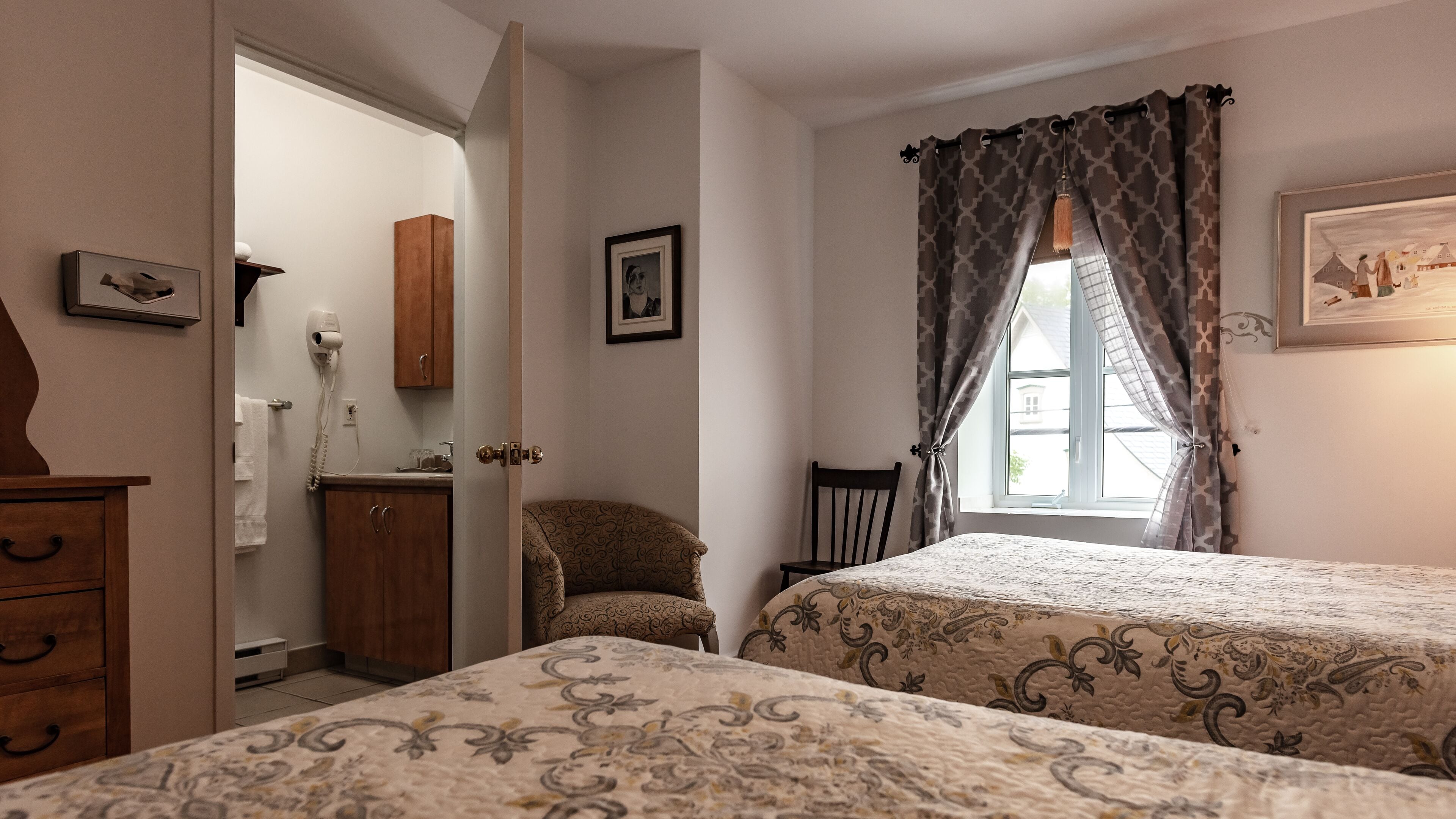 A bedroom with two beds, a wooden wardrobe, a chair, a window with patterned curtains, and a wall-mounted picture.