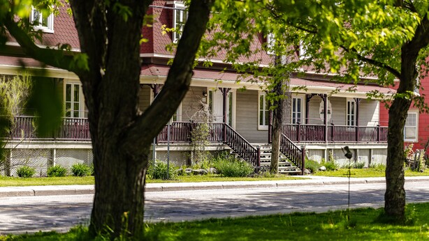 A row of two-story houses with a central staircase, surrounded by trees and a sidewalk. - Auberge Akamaraska (Kamouraska)