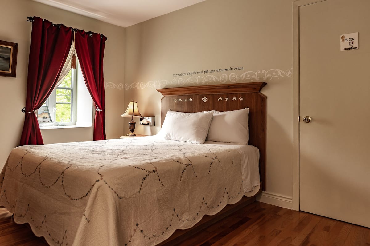 A bedroom with a wooden bed, a nightstand, a lamp, and a window with red curtains.