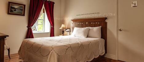 A bedroom with a wooden bed, a nightstand, a lamp, and a window with red curtains.
