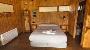 Habitación de cabaña de madera con una cama, un radiador, un estante con un tazón y una ventana con vistas.