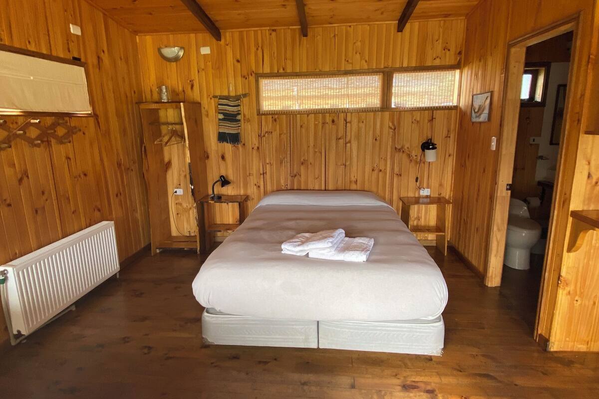 A wooden cabin room with a bed, a radiator, a shelf with a bowl, and a window with a view.