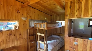 Interior de una cabaña de madera con literas, una mesa de comedor, sillas, una zona de cocina y un ventanal con vistas al exterior.
