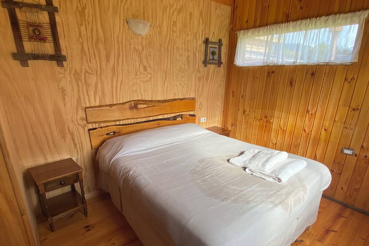 A wooden-paneled bedroom with a single bed, a nightstand, a wall-mounted light, and a window with sheer curtains.