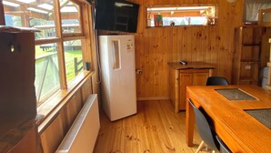 Interior de una cabaña de madera con zona de cocina, una mesa de comedor y ventana con vistas a un jardín.