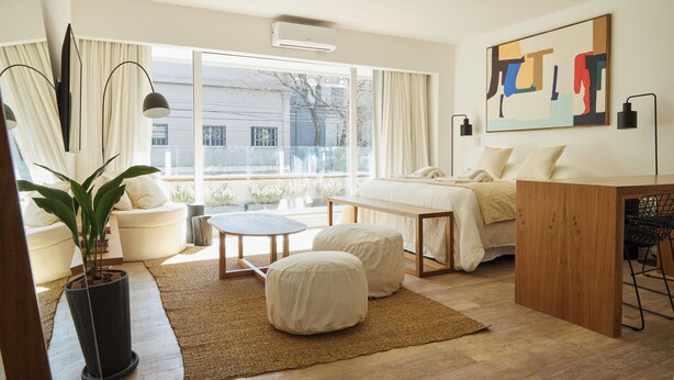 Een moderne slaapkamer met een groot raam, een bed, een nachtkastje, een potplant en een aan de muur bevestigd kunstwerk.