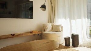 Een moderne woonkamer met een flatscreen televisie aan de muur, een houten bijzettafeltje, een ronde houten salontafel, een witte bank met kussens en een vloerlamp.