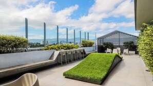 A rooftop terrace with a green artificial grass area, seating, and a view of the cityscape.