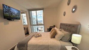 A bedroom with a large bed, a TV mounted on the wall, a window with a city view, and a bedside lamp.