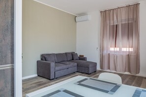 A living room with a gray sectional sofa, a glass coffee table, a white chair, a window with sheer curtains, and an air conditioning unit on the wall.