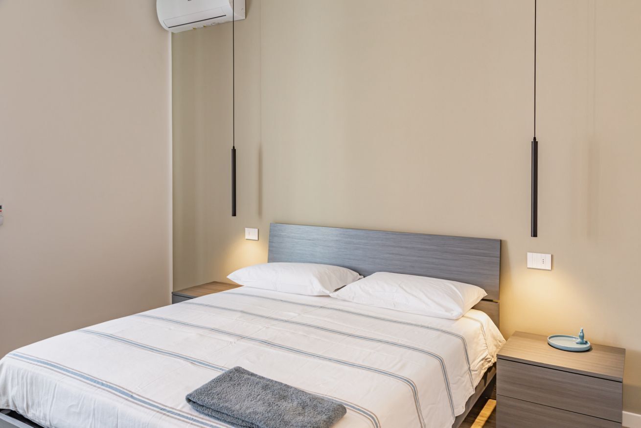 A bedroom with a bed, bedside tables, a wall-mounted air conditioner, and a nightstand with a lamp.