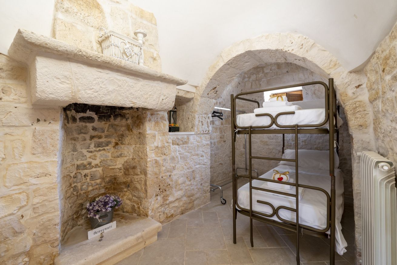 A bunk bed in a stone-walled room with a fireplace and a small shelf with a flower.