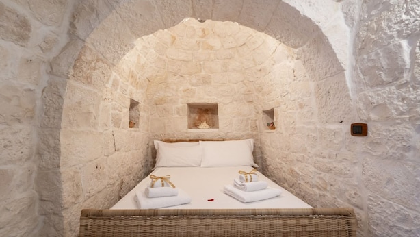 Una habitación de paredes de piedra con una cama, un pequeño rincón y una silla de mimbre.