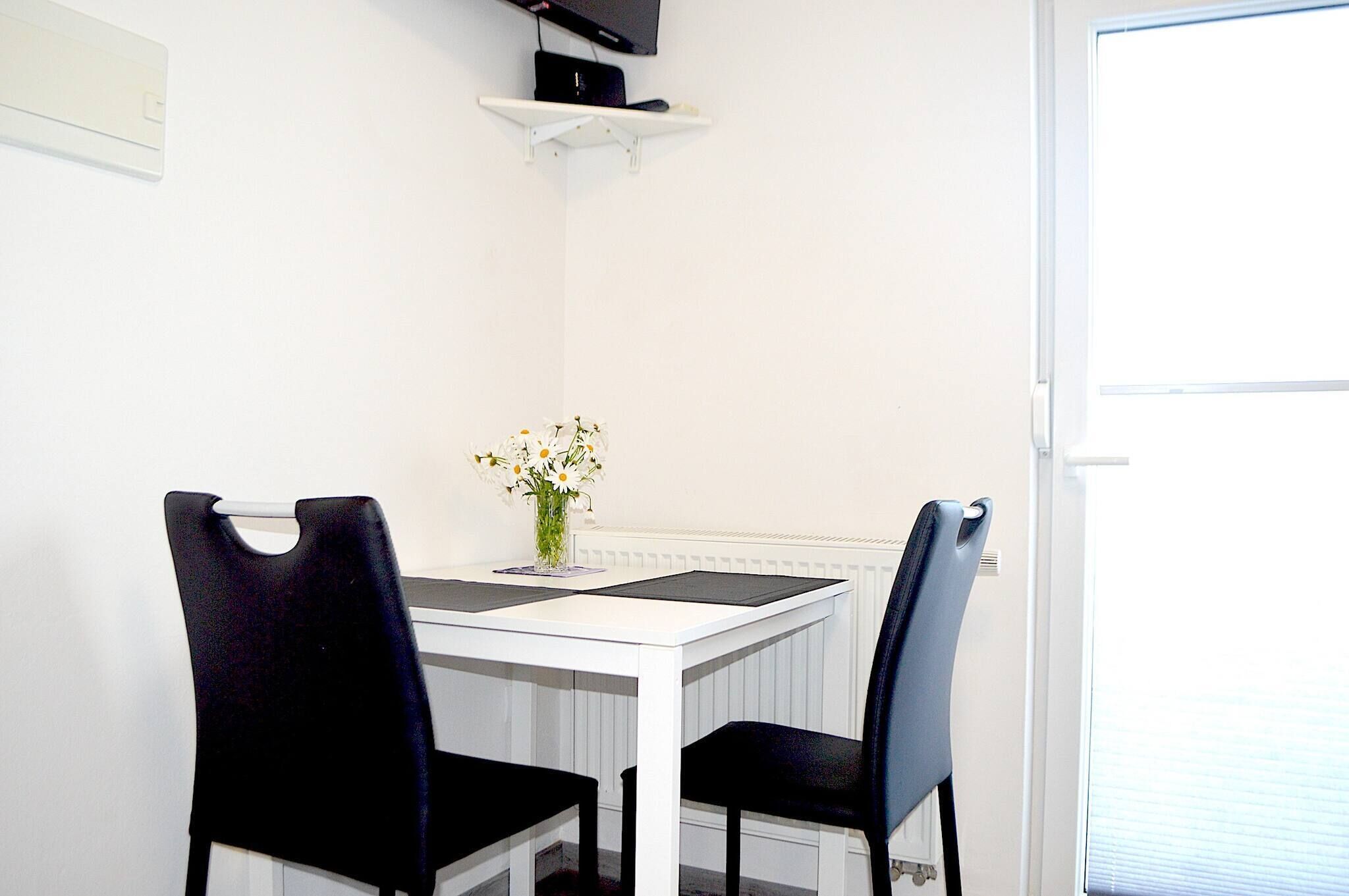 A white table with two black chairs, a vase with flowers, and a shelf with a black object.