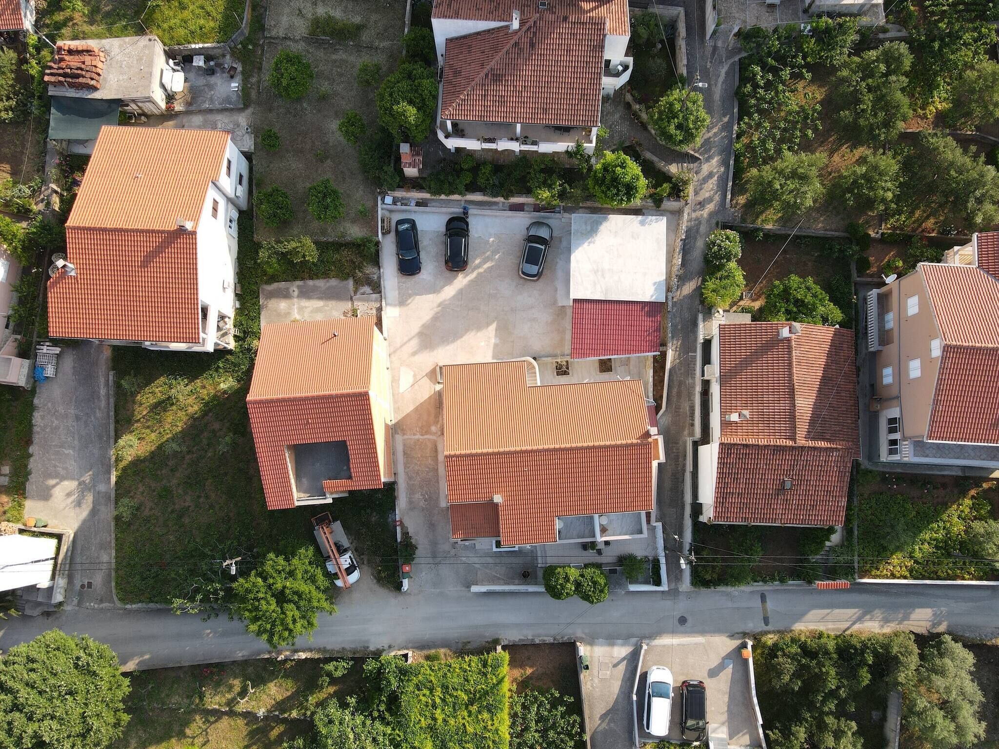 An aerial view of a residential area with houses featuring red-tiled roofs, a central parking area, and surrounding greenery.
