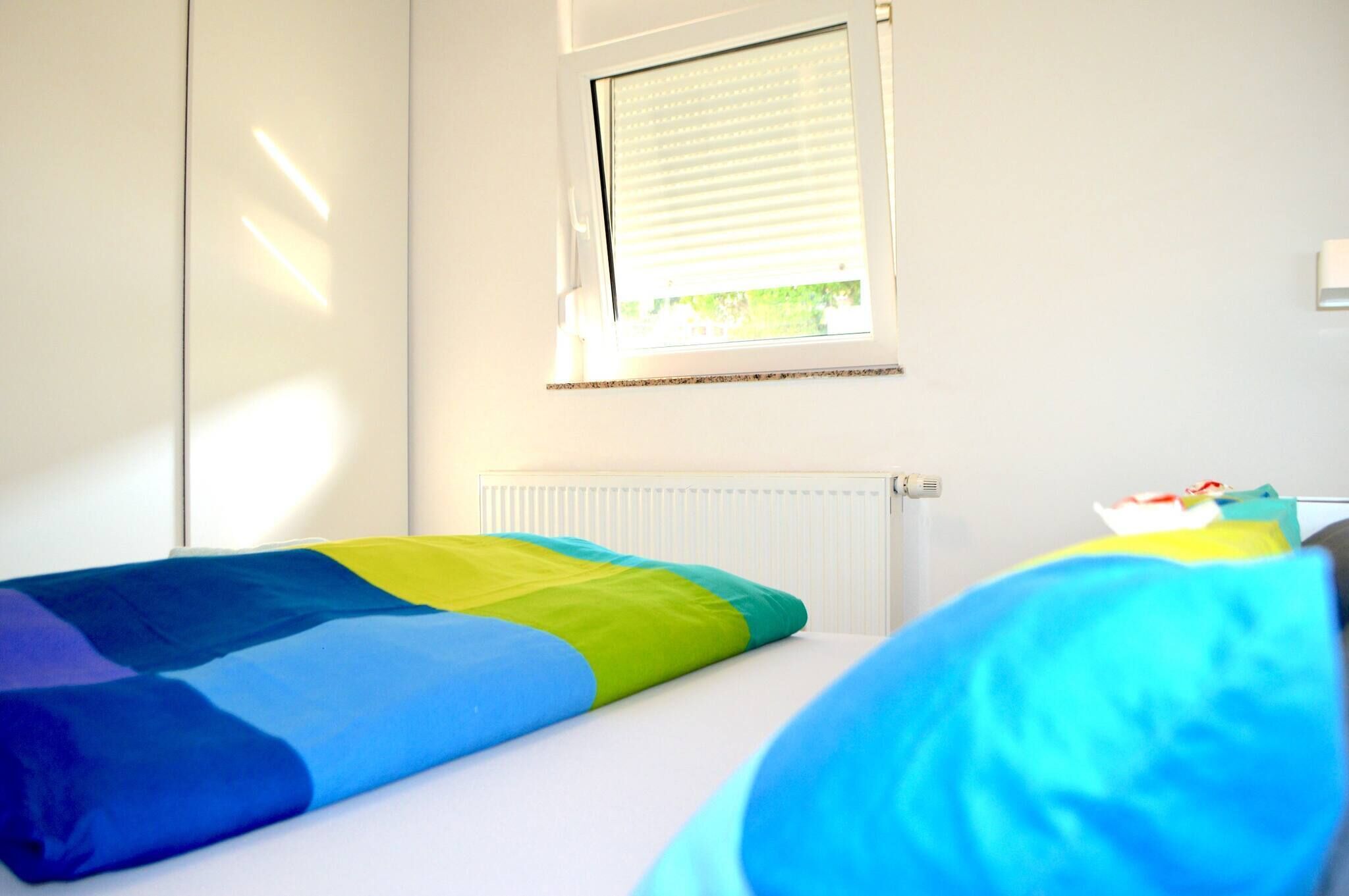 A room with a bed, a window with blinds, a radiator, and a wall-mounted light fixture.