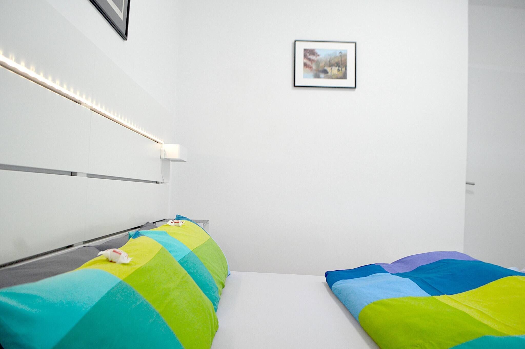 A modern hotel room with a bed, a bedside table, a lamp, and a framed picture on the wall.