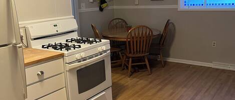 A kitchen with a stove, oven, and refrigerator. There is a dining table with chairs and a window with patterned curtains.