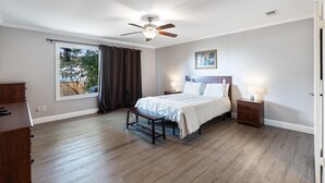 Een slaapkamer met een bed, nachtkastjes, een bankje, een plafondventilator, een raam met gordijnen en uitzicht op bomen.