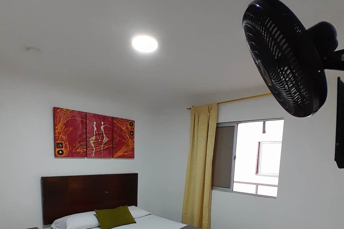 A neatly arranged bedroom with a bed, a bedside table, a wall-mounted fan, a window with curtains, and a painting on the wall.